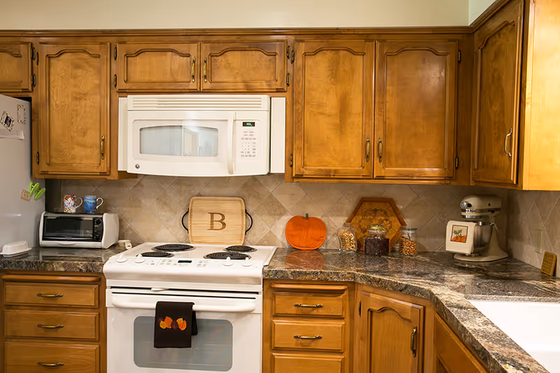 A kitchen with wooden cabinets, a white electric stove with a black towel hanging on the handle, a white microwave above the stove, a toaster oven on the counter, and various kitchen items including a pumpkin decoration, jars, and a stand mixer on the granite countertop.