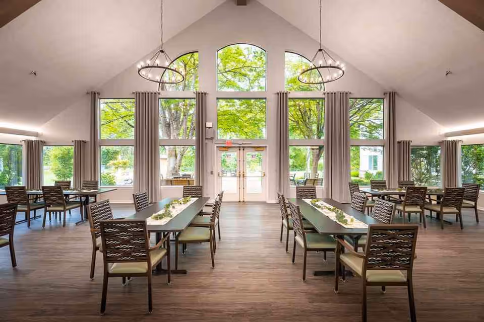 Bright dining hall with long tables, chairs, and large arched windows overlooking greenery.