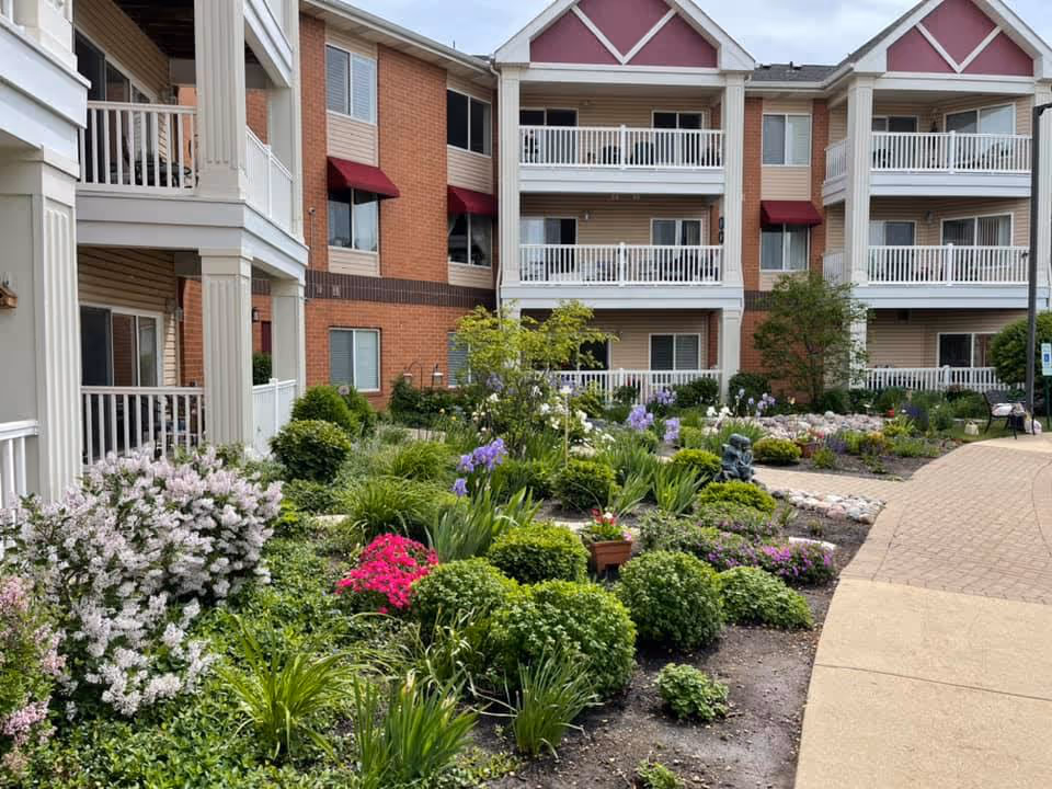 Three-story senior living building with balconies surrounding a colorful landscaped courtyard garden.