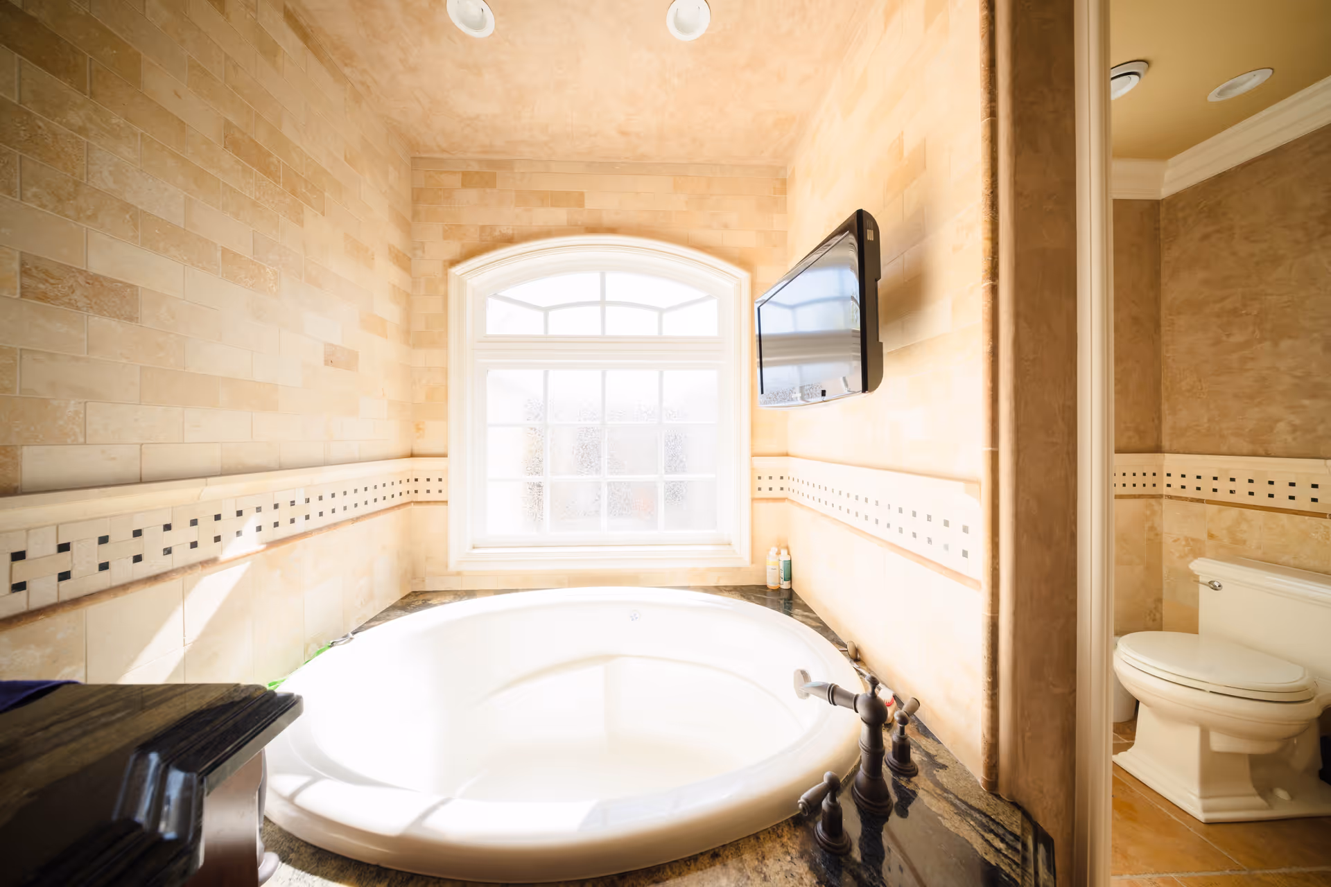 Bright bathroom with a circular tub, window, tiled walls, and a wall-mounted TV.
