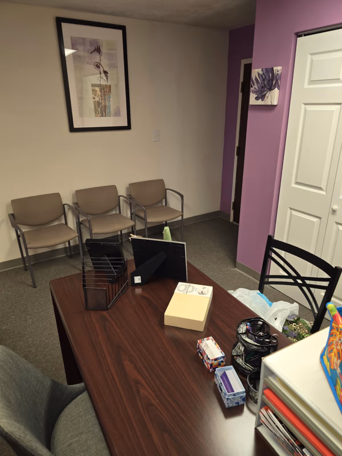 Office desk in a waiting area with four chairs along a beige wall and purple accents.