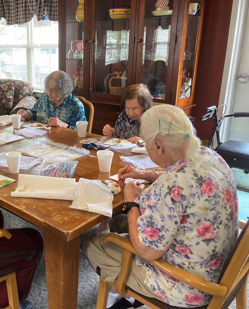 Three elderly women sitting around a wooden table engaged in painting activities. The table has art supplies including paint palettes, colored pencils, and paper towels. Behind them is a wooden cabinet with glass doors displaying glassware and decorative items. The room has a window with patterned curtains letting in natural light.