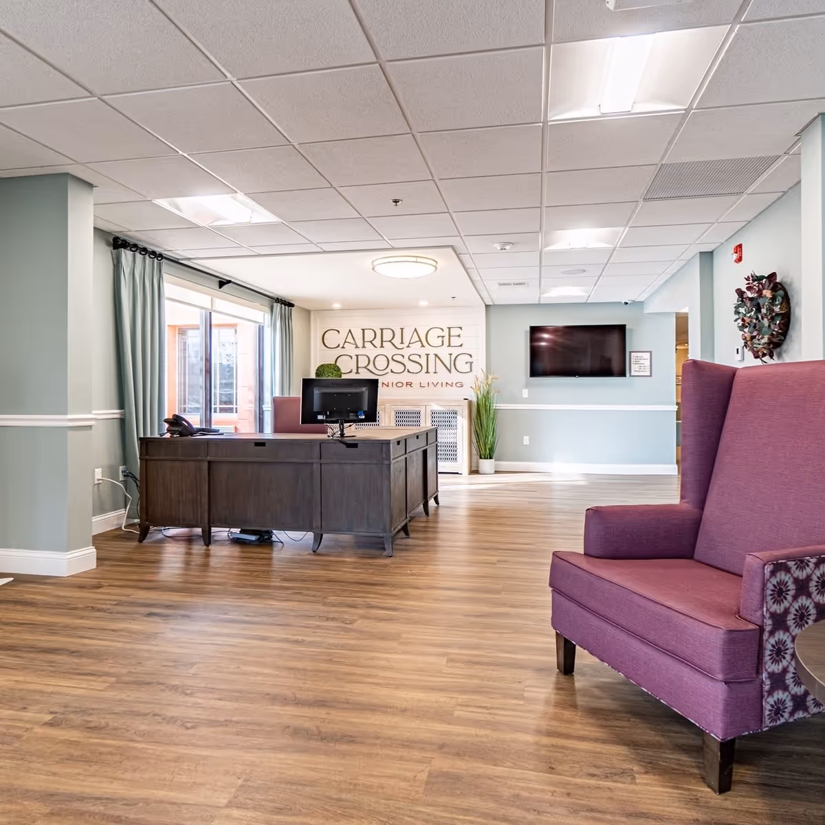 Interior view of a senior living facility reception area with a wooden desk, computer monitor, and a purple upholstered armchair. The walls are painted light blue with white trim, and there is a large window with curtains on the left. A flat-screen TV is mounted on the wall, and a decorative plant and wall art are visible. The sign on the back wall reads 'Carriage Crossing Senior Living.'
