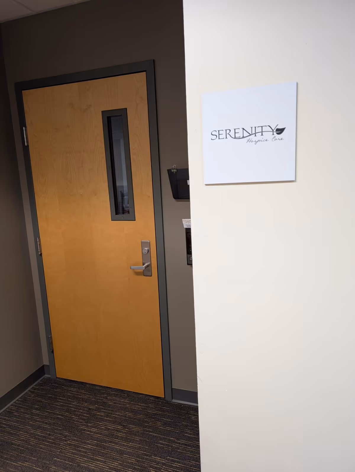 Wooden interior door in a hallway beside a Serenity Hospice Care sign on the wall.