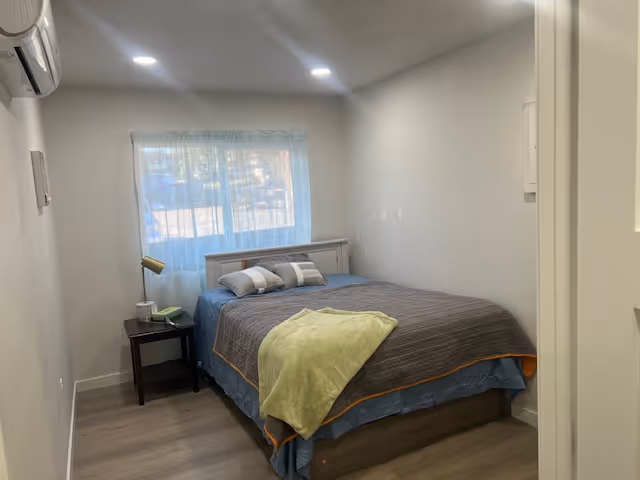 Small bedroom with a bed, nightstand, lamp, and window in white walls.