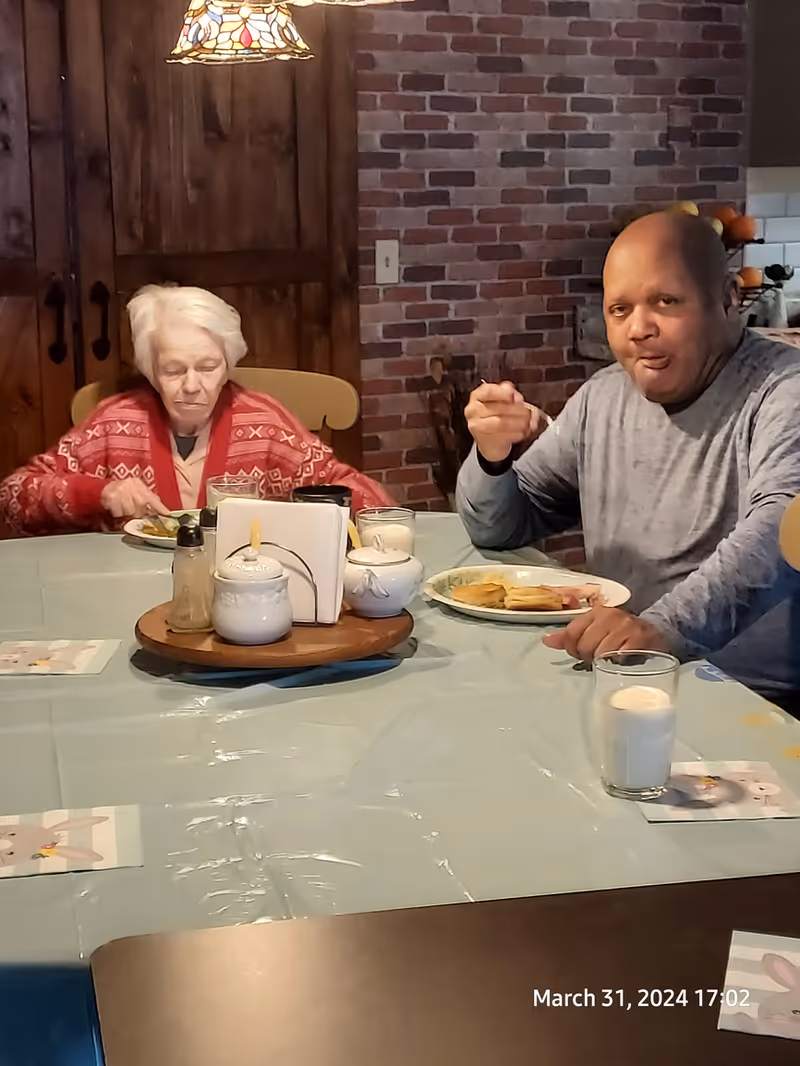 Two elderly individuals sitting at a dining table eating a meal. The table is covered with a light blue tablecloth and has a round wooden tray with condiments and napkins in the center. The background features a brick wall and wooden cabinets. One person is wearing a red patterned sweater and the other is in a gray long-sleeve shirt. A glass of milk is visible on the table.