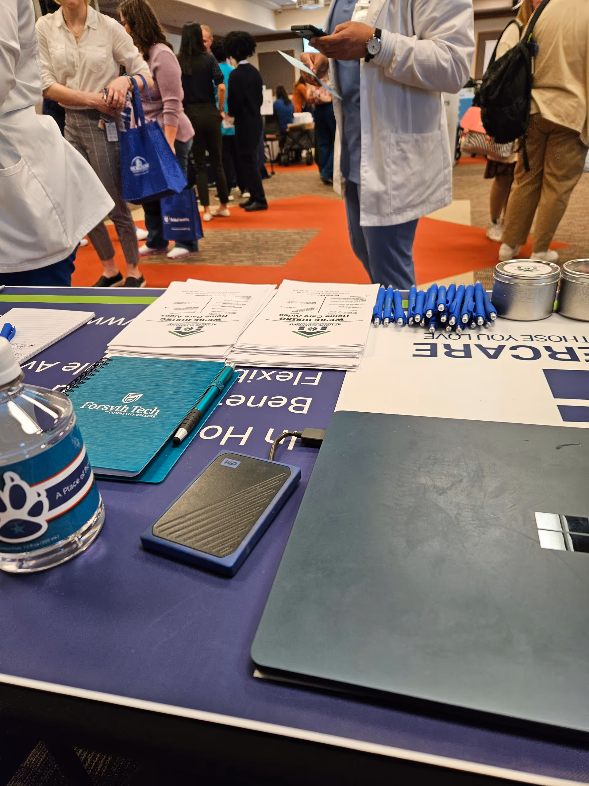 Indoor registration desk with brochures, pens, a laptop, and attendees at an eldercare event.