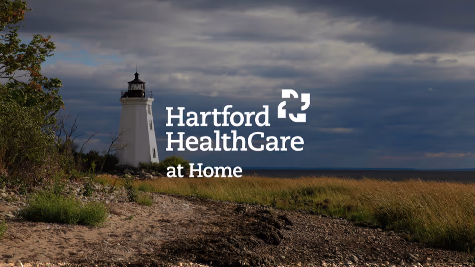 Lighthouse by a rocky shore with grasses and a Hartford HealthCare at Home logo overlay.