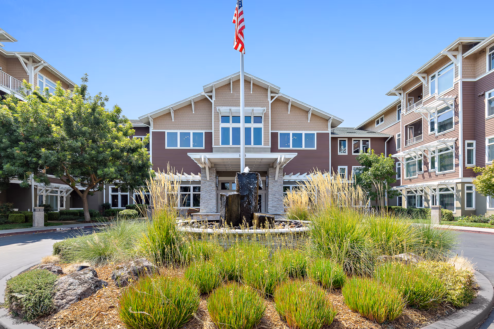 Front entrance of a multi-story retirement community building with a central fountain, landscaped roundabout, and an American flag.