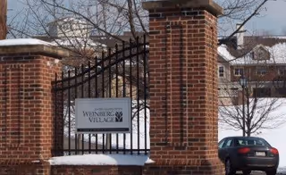 Brick entrance gate with a Weinberg Village sign, snow-covered ground, residential buildings in background.
