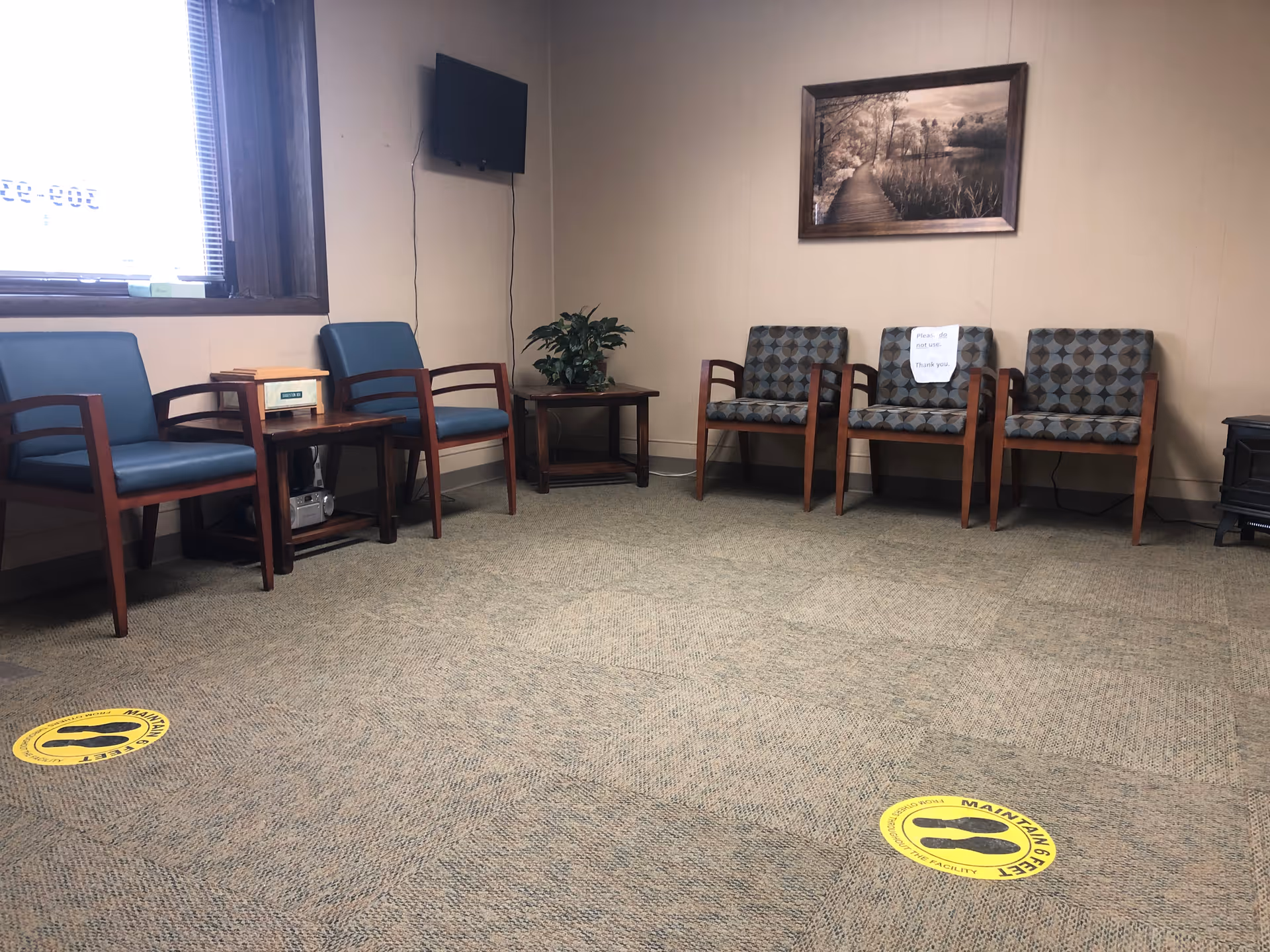 Waiting room with blue chairs, wall art, a plant, TV, and yellow social distancing markers.
