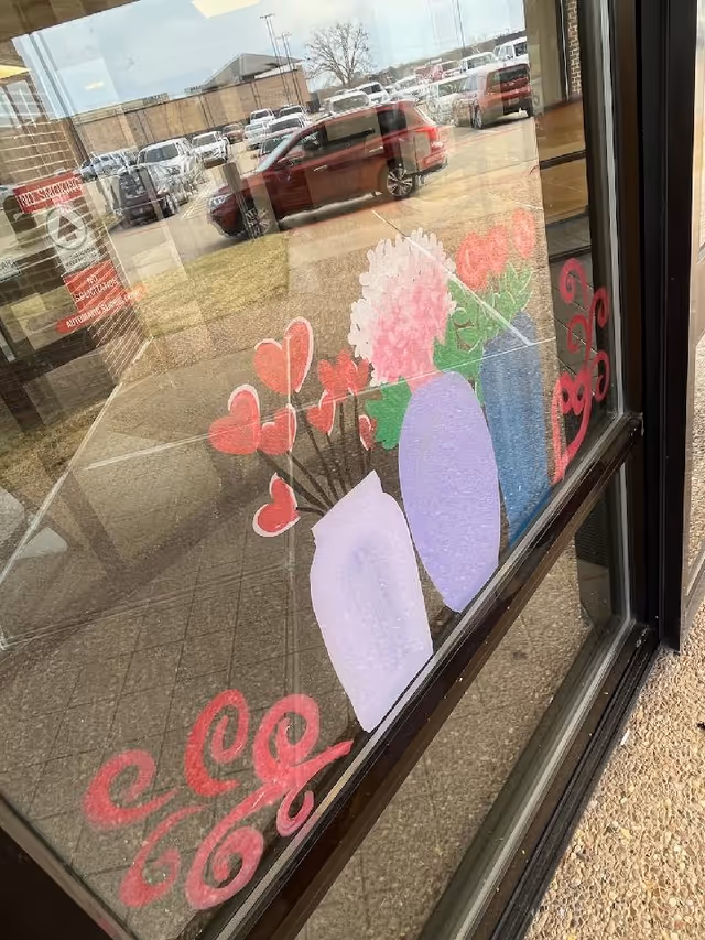 Storefront window with decorative flower and heart decals; parking lot reflected outside.