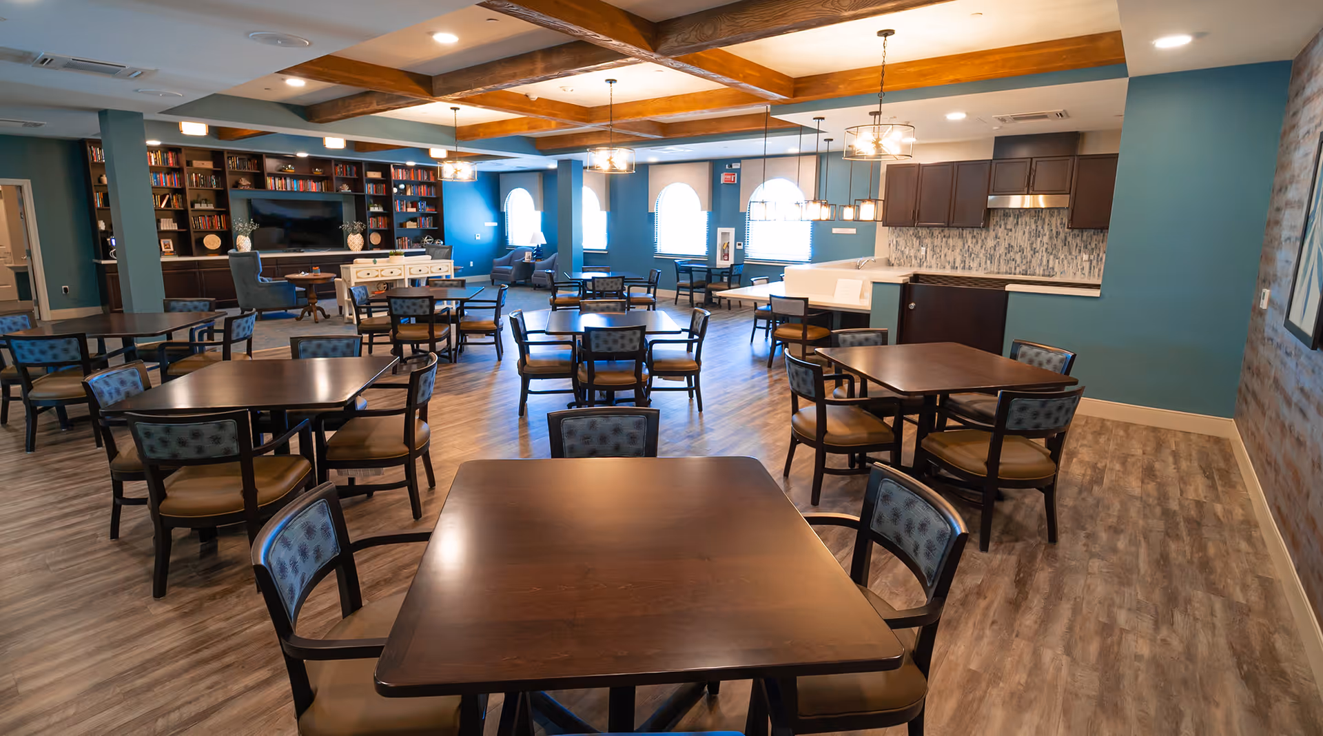 Spacious senior living dining and common area with multiple tables and chairs, a kitchenette, and a lounge with bookshelves.