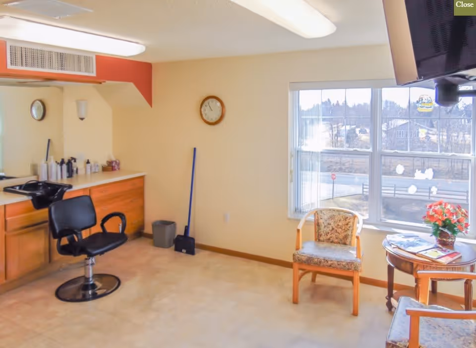 A small salon-style interior with a styling chair and sink, cabinets, and chairs beside a large window.