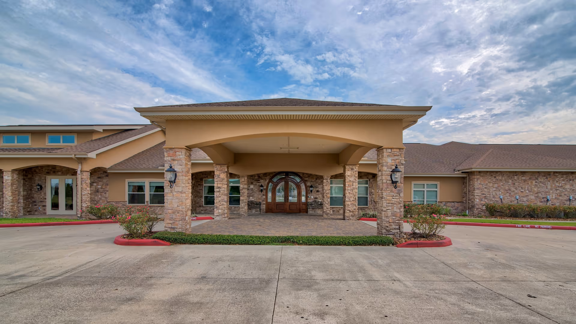 Front entrance of a single-story assisted living building with a covered porte-cochere, stone columns and arched double doors under a partly cloudy sky.