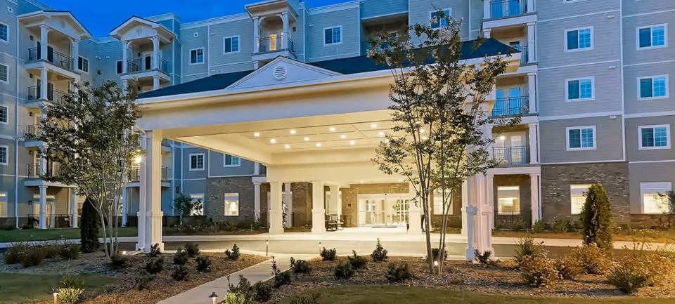 Entrance of a multi-story senior living facility named Towne Club Windermere at dusk, featuring a covered drop-off area with columns, landscaped garden beds, and illuminated windows.