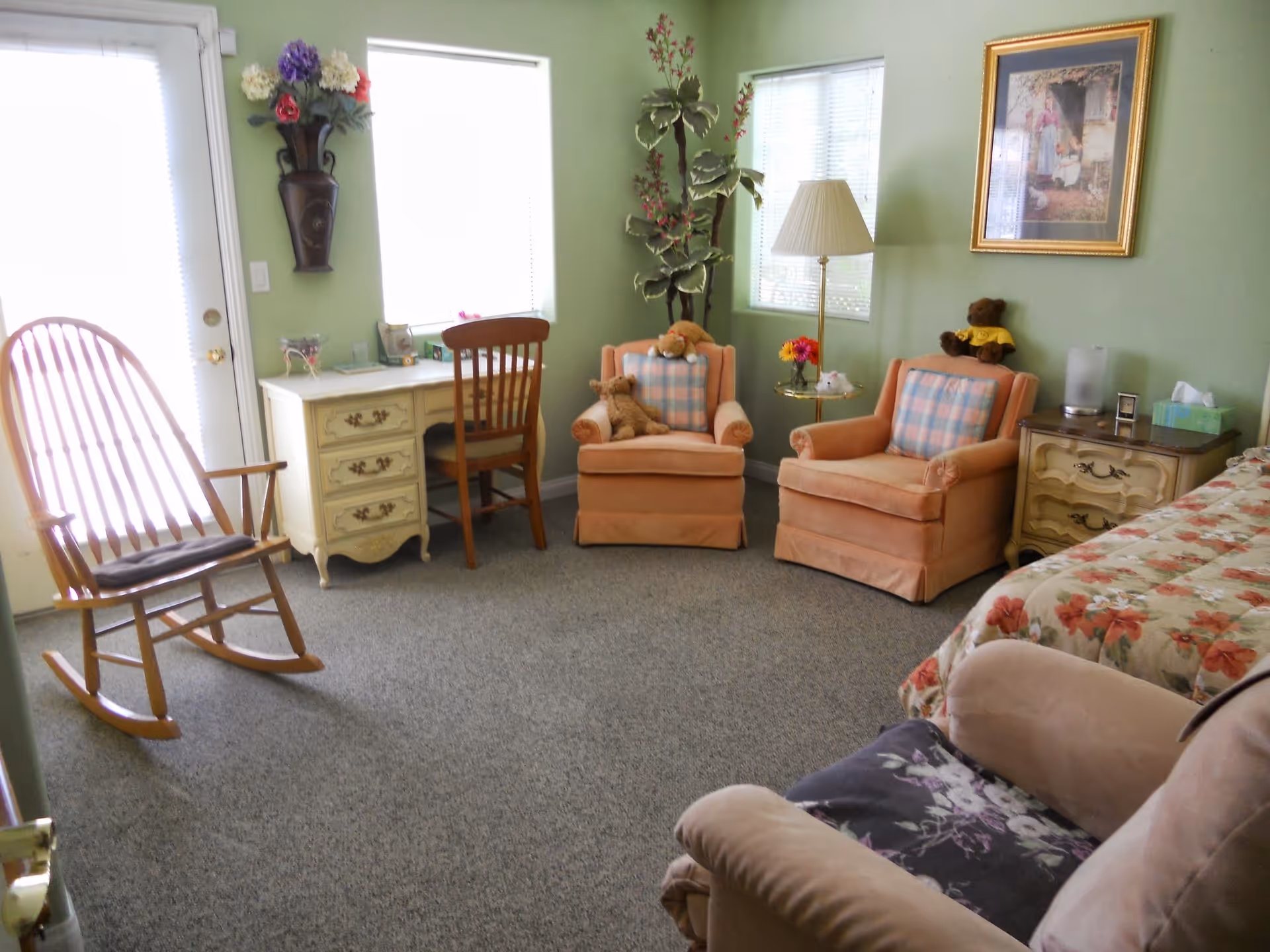 A cozy room with green walls featuring a wooden rocking chair, a small desk with a wooden chair, two peach-colored armchairs with plaid cushions and teddy bears, a floor lamp, a side table with flowers and a tissue box, a bed with floral bedding, and a framed picture on the wall.