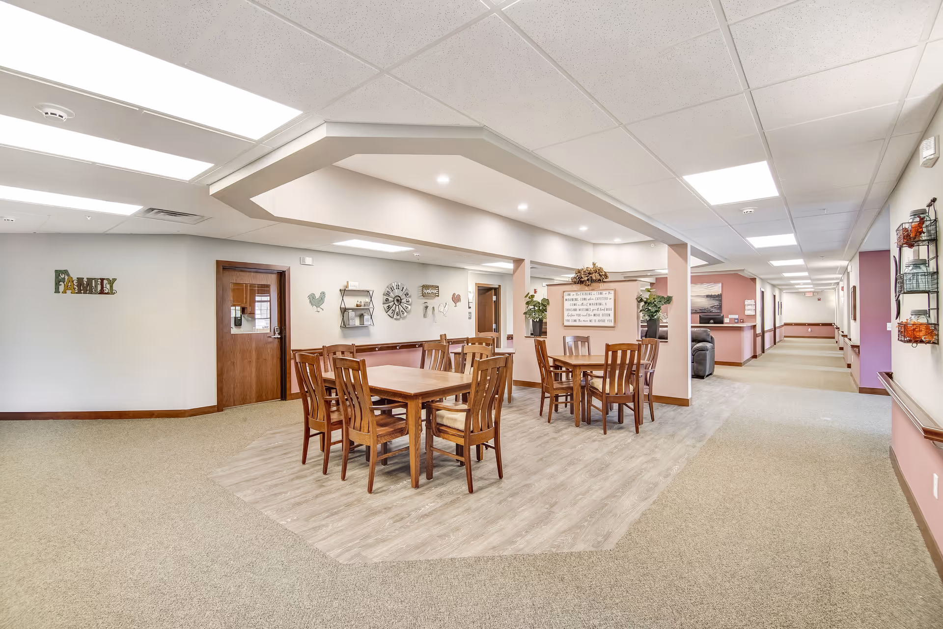 Open communal dining area in a senior living facility with wooden tables and chairs under recessed lighting and a long hallway to the right.