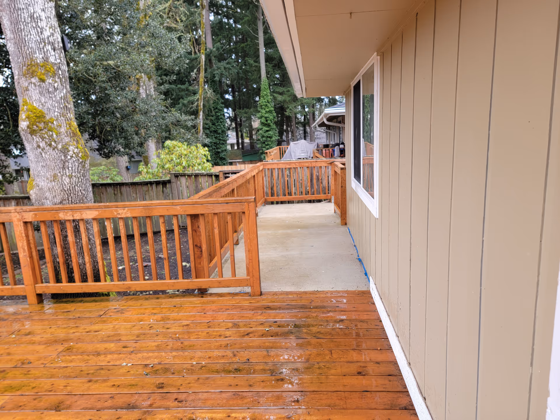 An outdoor wooden deck with railing beside a beige building and surrounding trees.