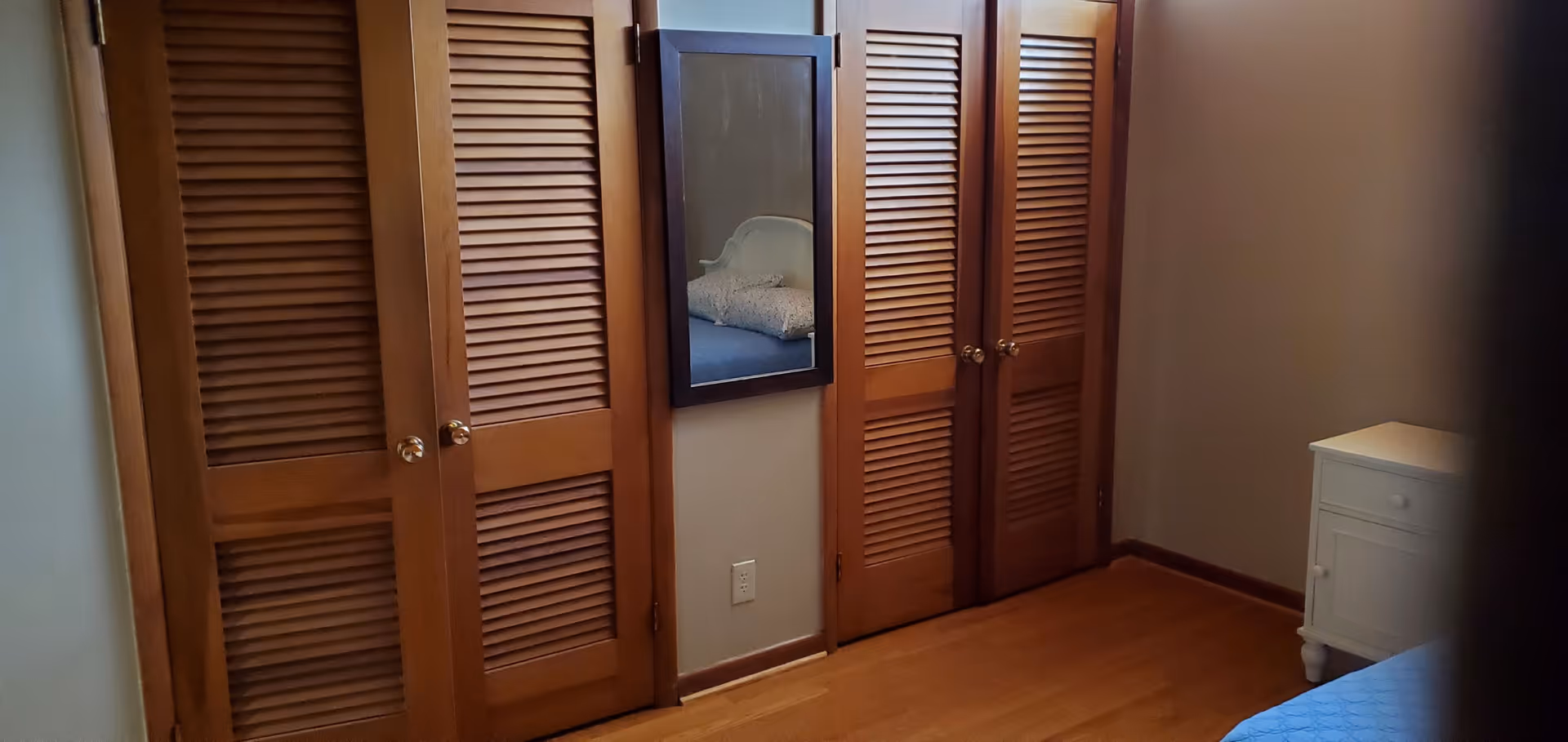 Bedroom with wooden louvered closet doors, wall mirror, blue bedding, and white nightstand.