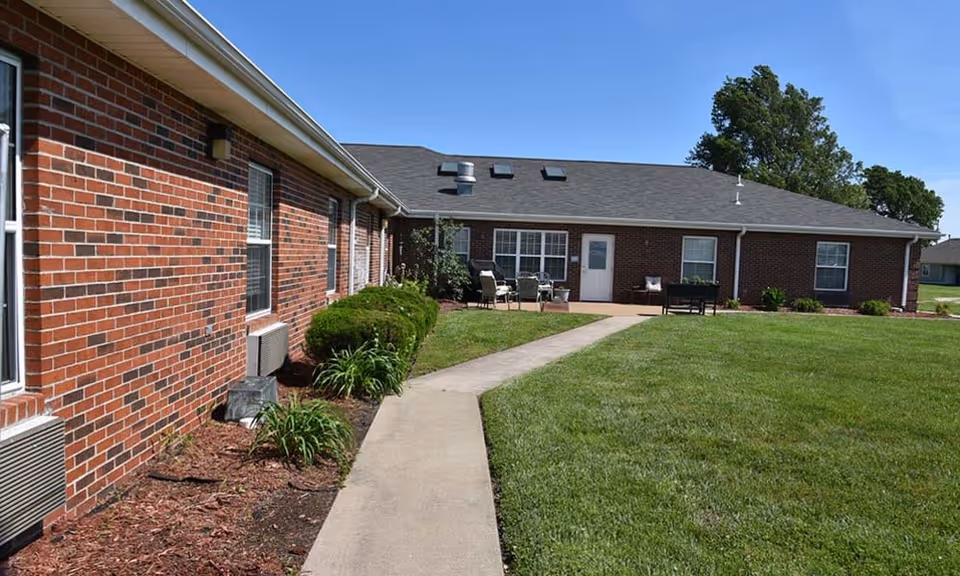 Single-story red brick assisted living building exterior with a concrete path leading to a patio area and a green lawn.