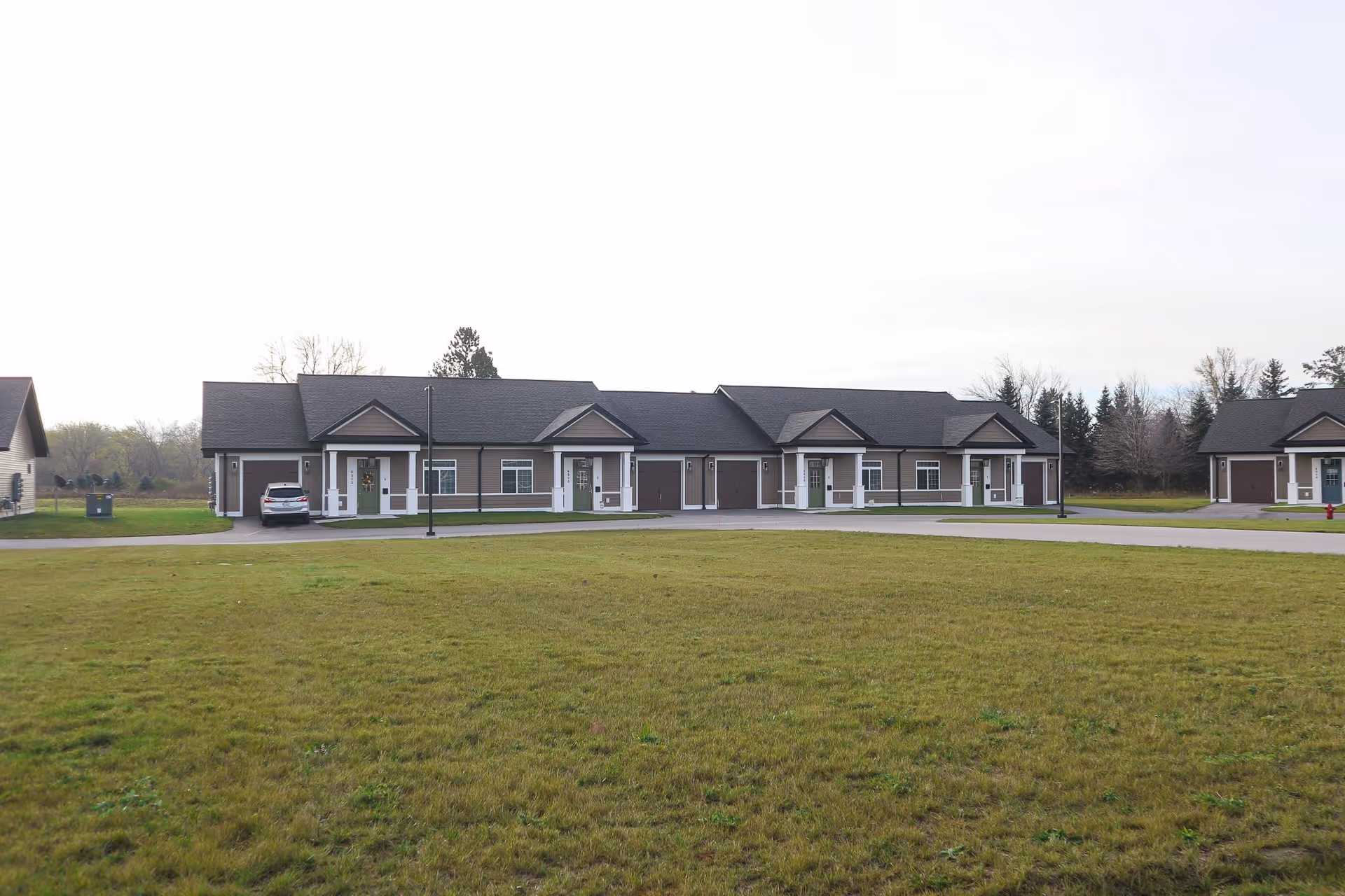 Single-story row of connected residential units with garages facing a large grassy lawn under an overcast sky.