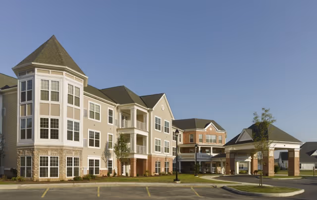 Three-story brick-and-siding senior living building with a turret and covered porte-cochère entrance under a clear blue sky.