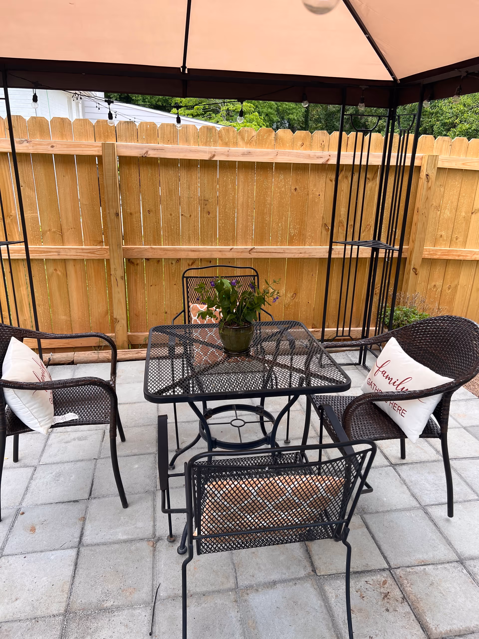 Outdoor patio area with a black metal table and four matching chairs, two of which have white cushions with red text. A small potted plant is placed on the table. The area is covered by a canopy and enclosed by a wooden fence.