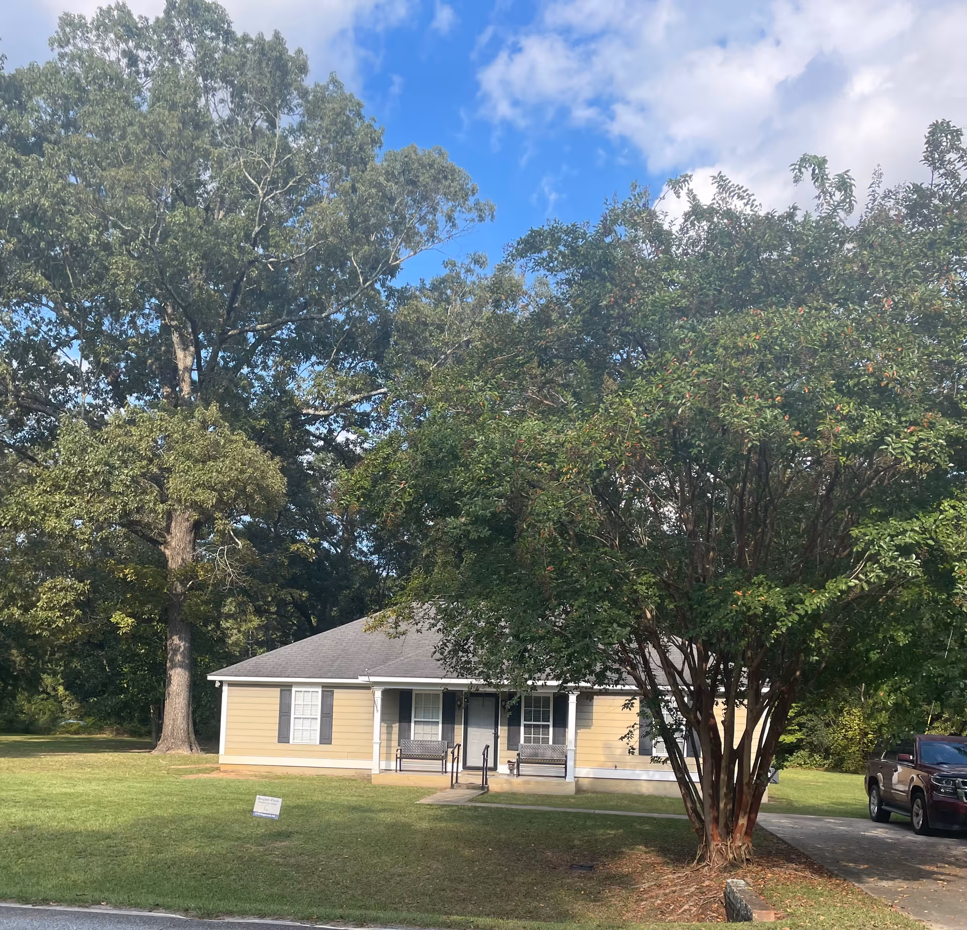Small yellow building with a front porch and benches, surrounded by large trees.