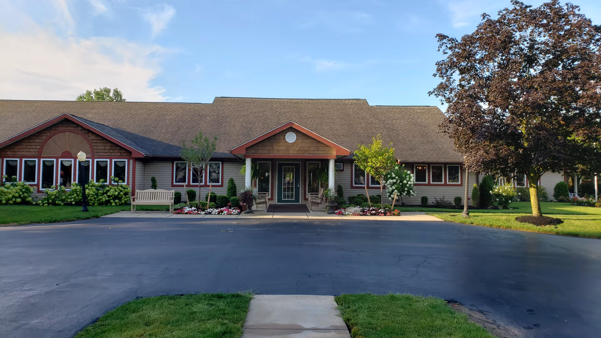 Front entrance of a single-story senior living building with a paved circular driveway, landscaped lawn, trees, benches, and flowering beds under a blue sky.