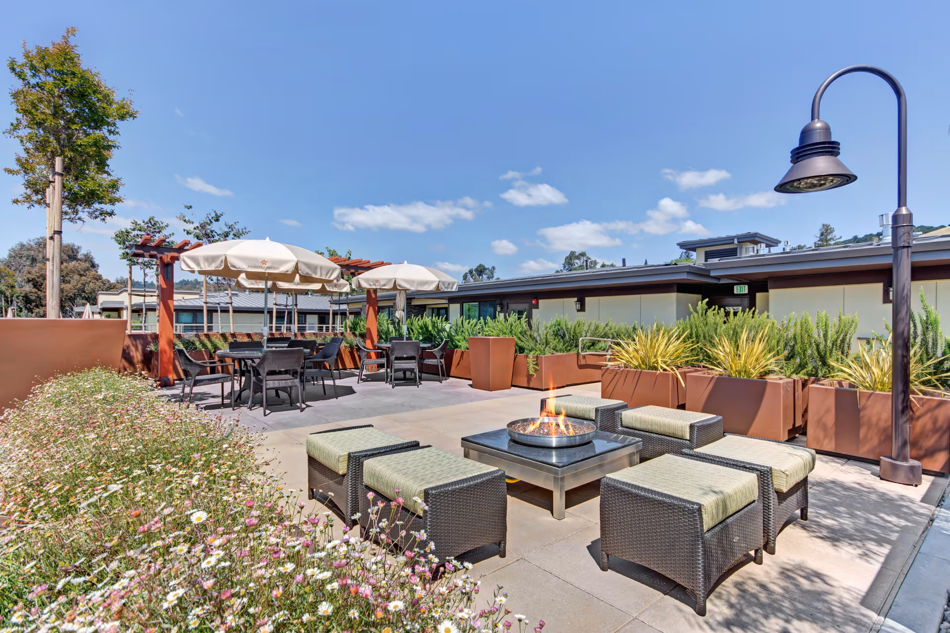 Outdoor patio area at Merrill Gardens at Lafayette featuring cushioned wicker seating around a fire pit, tables with umbrellas under a wooden pergola, potted plants, and a tall street lamp under a clear blue sky.