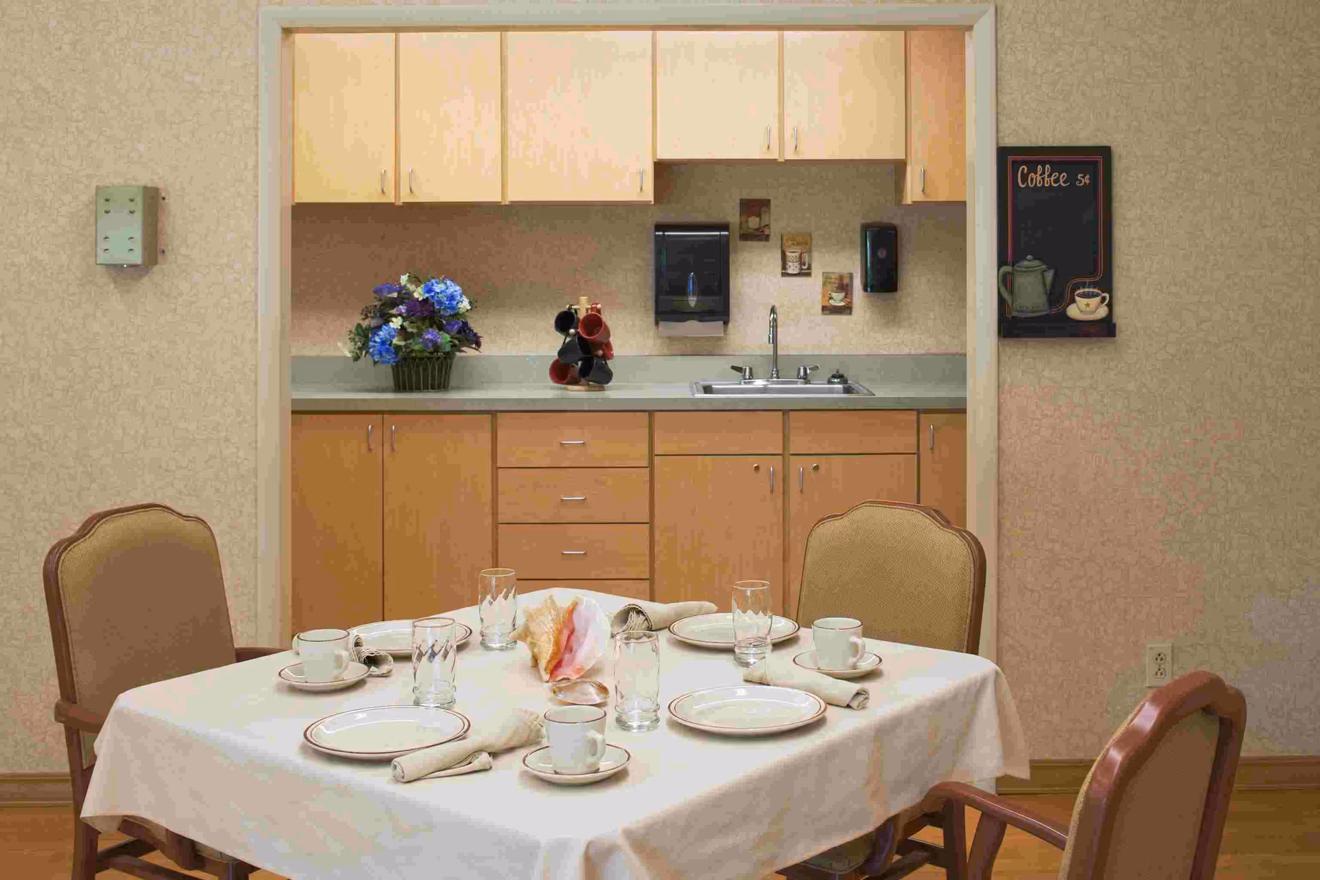 A dining room scene with a table set for meals and a kitchenette in the background.