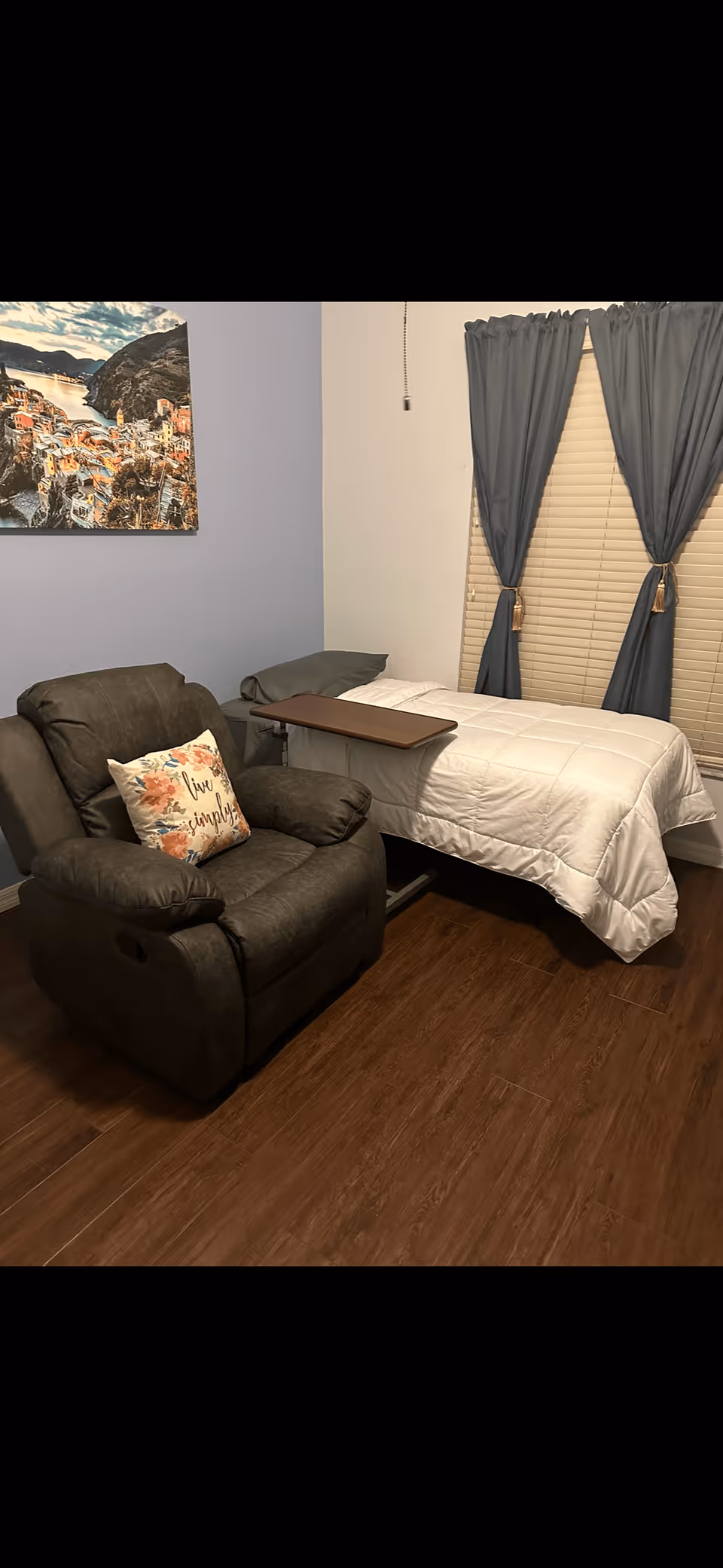 A small bedroom with a reclining armchair next to a single bed with a bedside tray, blue curtains, and wall art.
