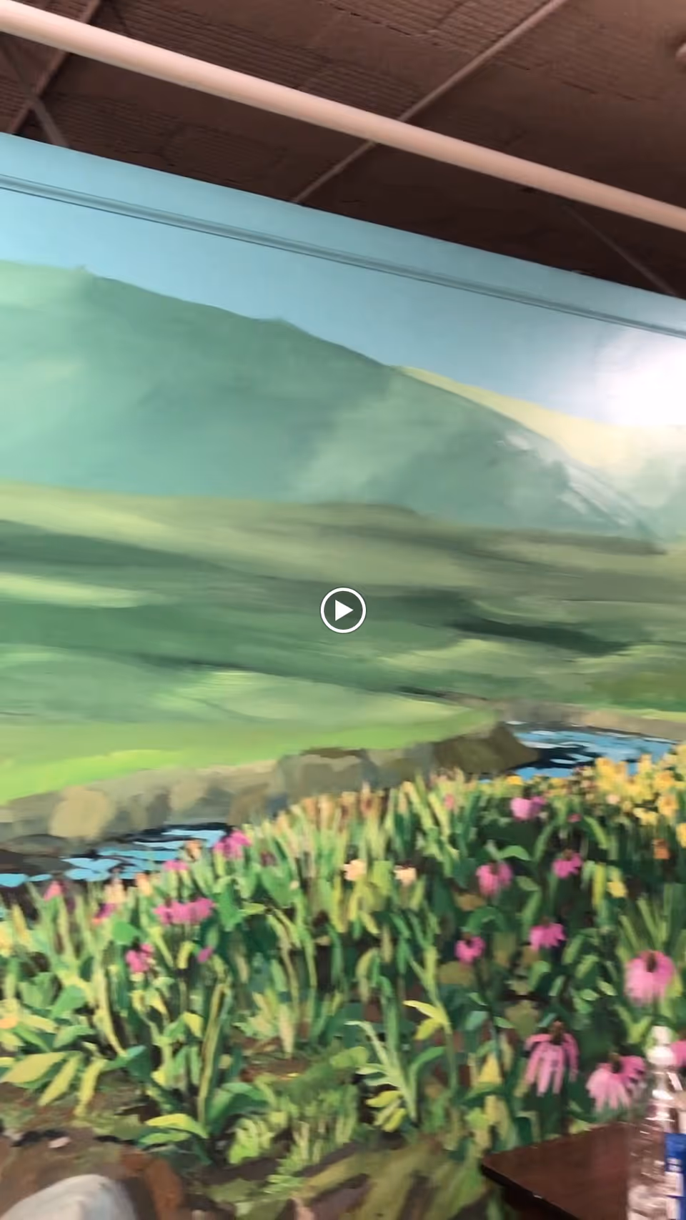 A mural painting depicting a natural landscape with green hills, a flowing river, and a field of pink and yellow flowers inside an indoor space with a visible ceiling structure.