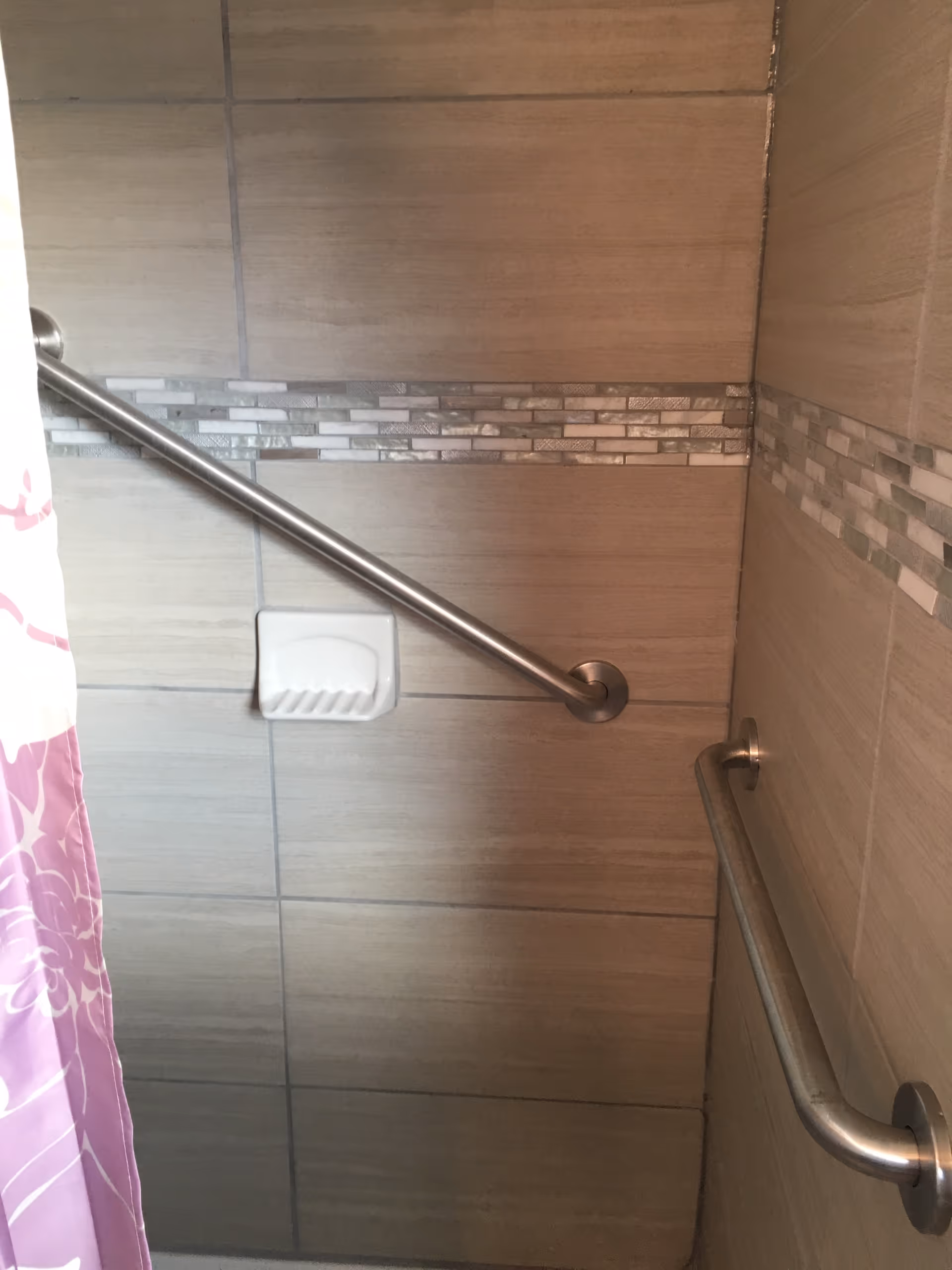 Shower area with beige tiled walls featuring a horizontal decorative strip of small rectangular tiles. There are two stainless steel grab bars mounted on the walls and a white soap dish. A pink and white floral shower curtain is partially visible on the left side.