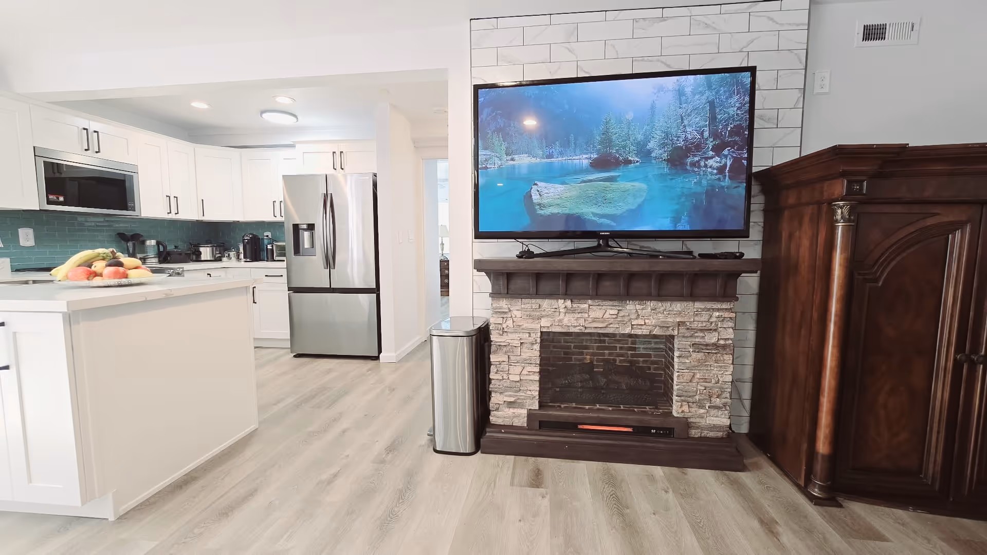 Open-concept kitchen and living area with white cabinets, island, stainless appliances, fireplace, and TV.