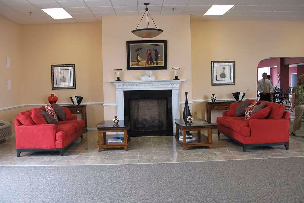 A cozy living room area with two red sofas facing each other across two wooden coffee tables. A white fireplace with decorative items and a framed painting above it is centered on the back wall. There are side tables with vases and framed pictures on the walls. In the background, a few people are walking through an adjacent room.