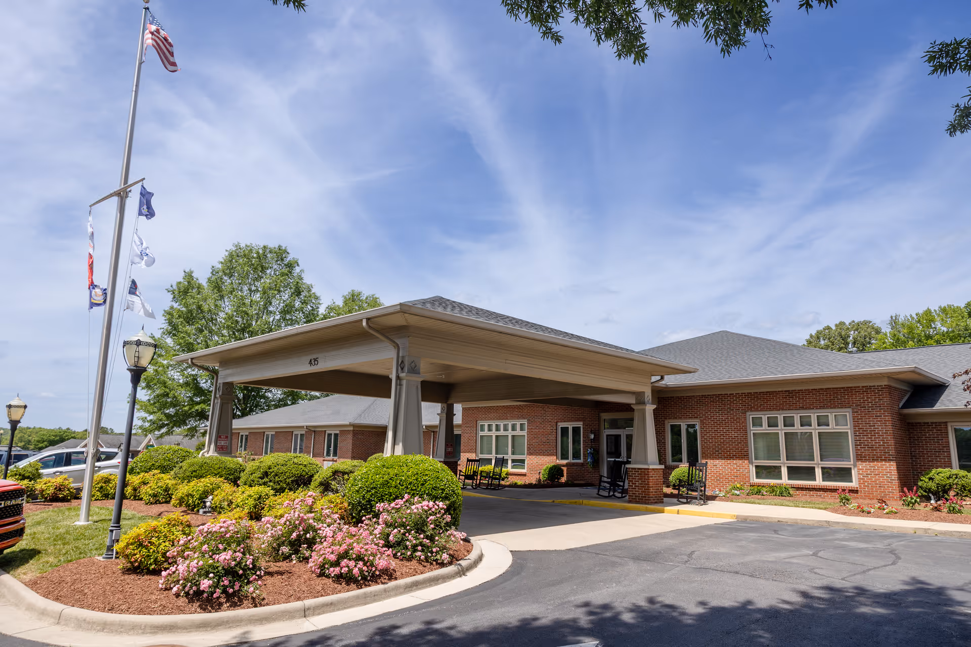 Brick senior living facility front entrance with a covered porte-cochere, flags, and landscaped shrubs.