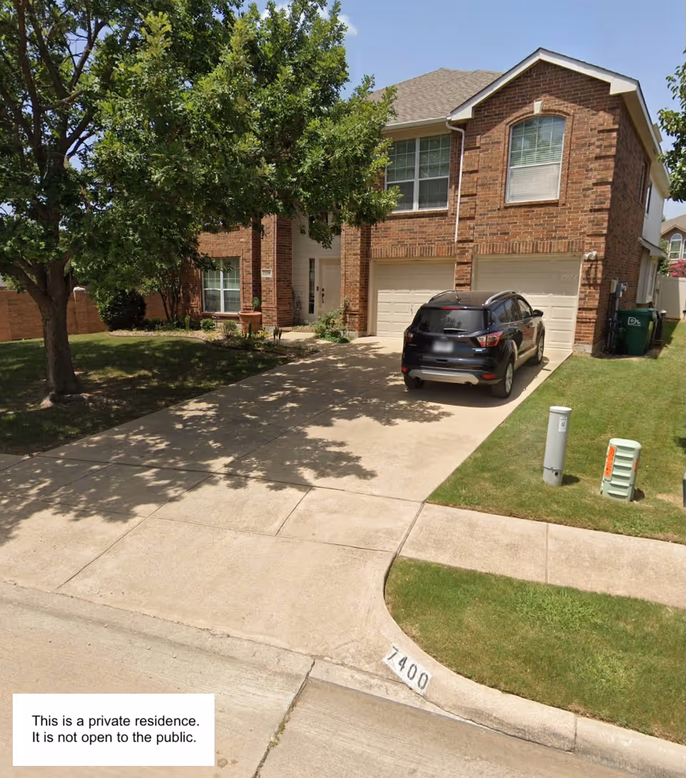 A brick two-story house with a driveway, parked car, and mature trees in front yard.