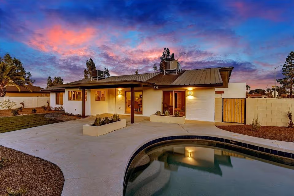 Exterior view of a single-story building at sunset with a swimming pool in the foreground, a concrete patio, and landscaped yard with plants and trees. The sky is colorful with shades of blue, purple, and pink.