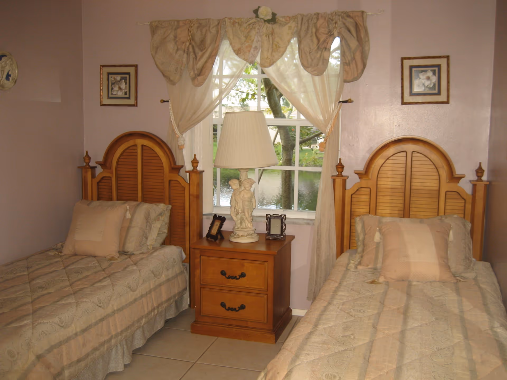 Two twin beds with wooden headboards flanking a nightstand and lamp in a softly lit bedroom with a window.
