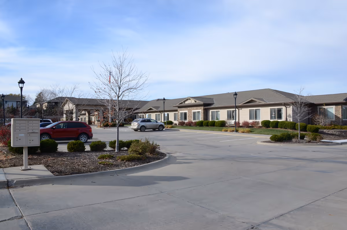 Exterior view of Cedar Creek of High Plains facility showing a single-story building with beige siding and stone accents, a parking lot with a few cars, leafless trees, and a mailbox cluster under a partly cloudy sky.