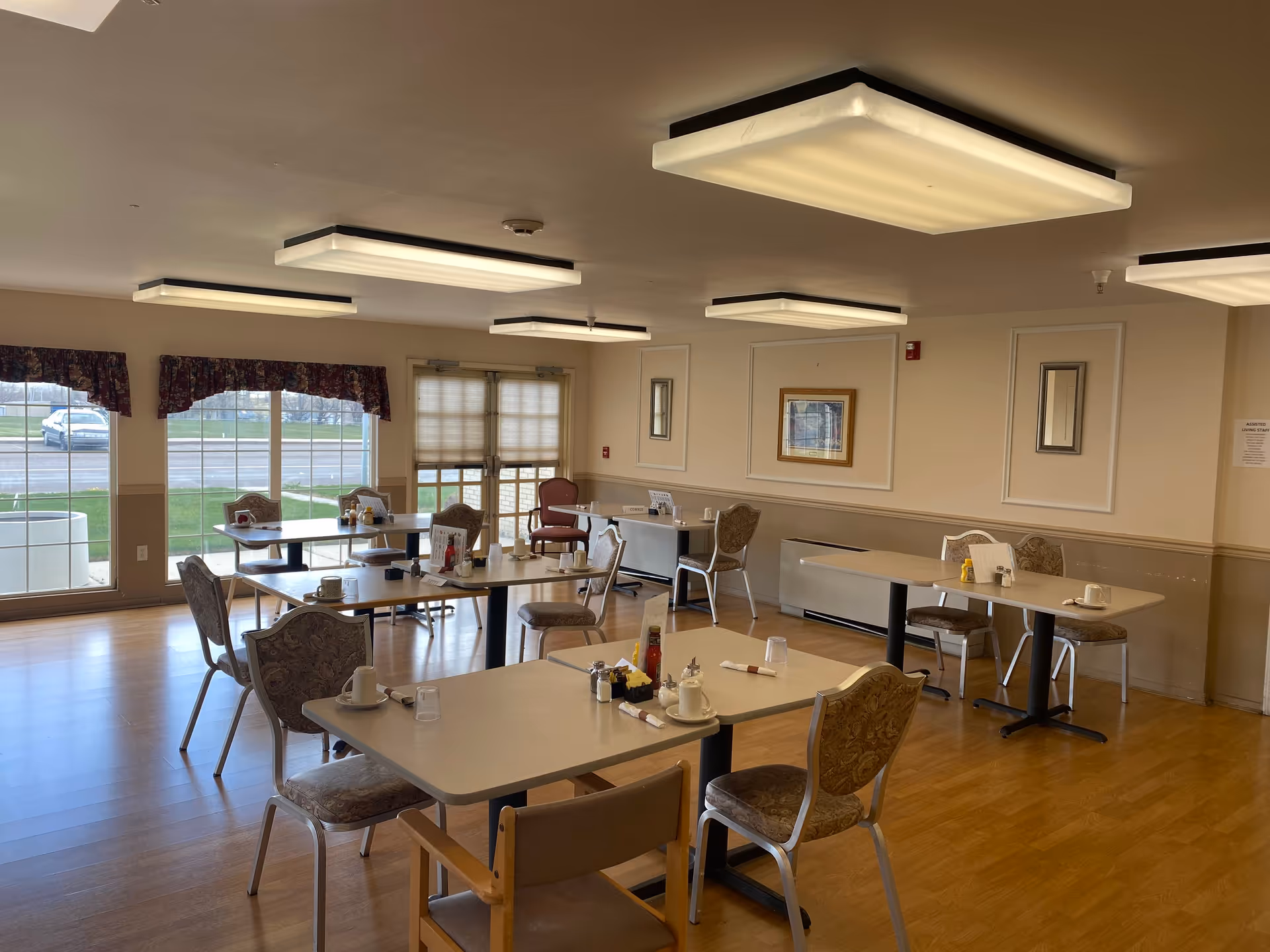Bright dining room with multiple tables, chairs, and place settings in a care facility.