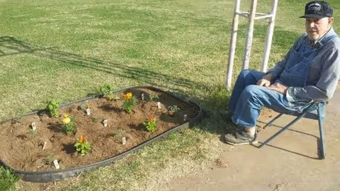 An elderly man sits outdoors on a chair beside a small garden bed.