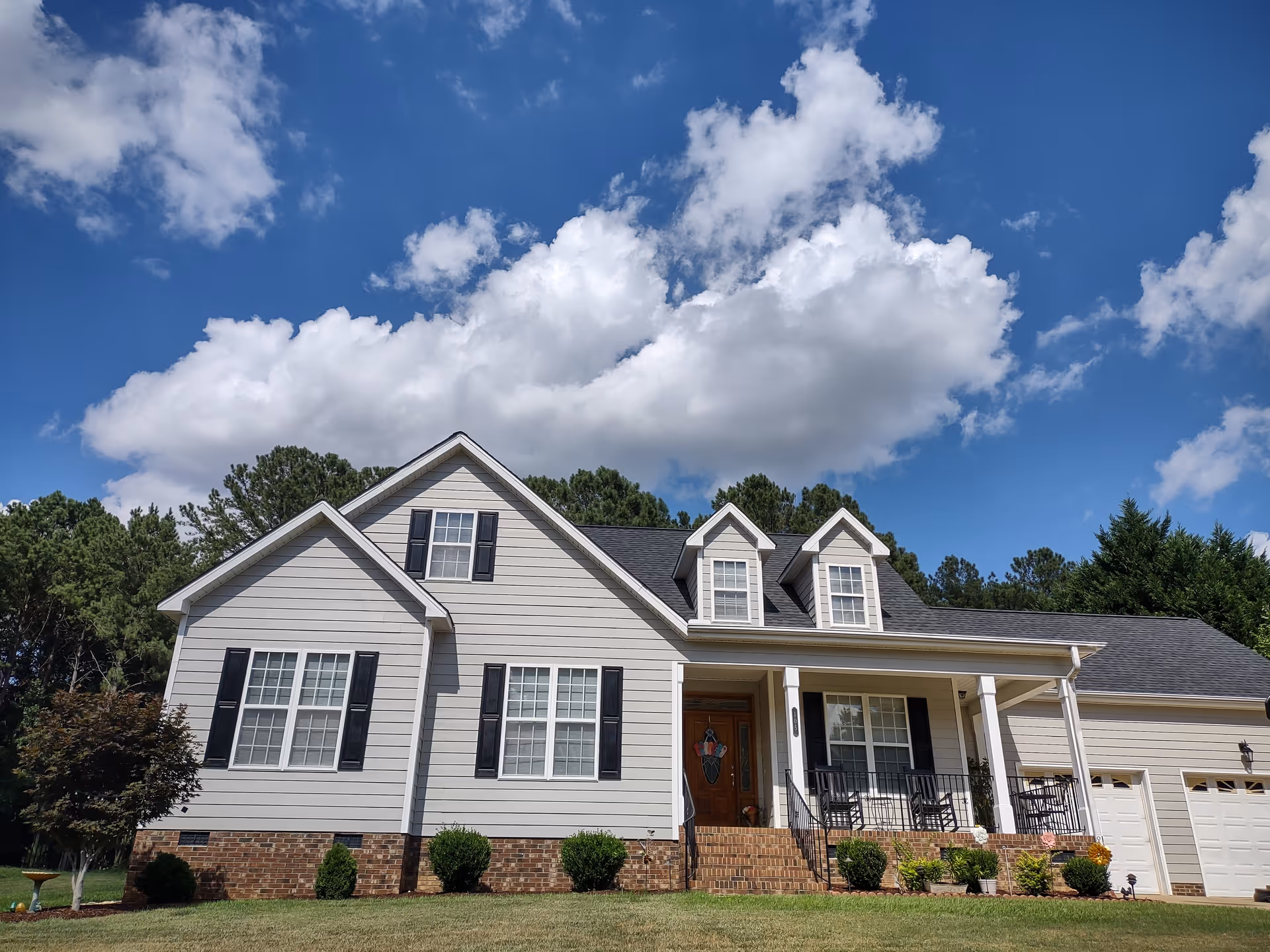 A two-story beige house with front porch, shutters, dormers, and a manicured lawn.