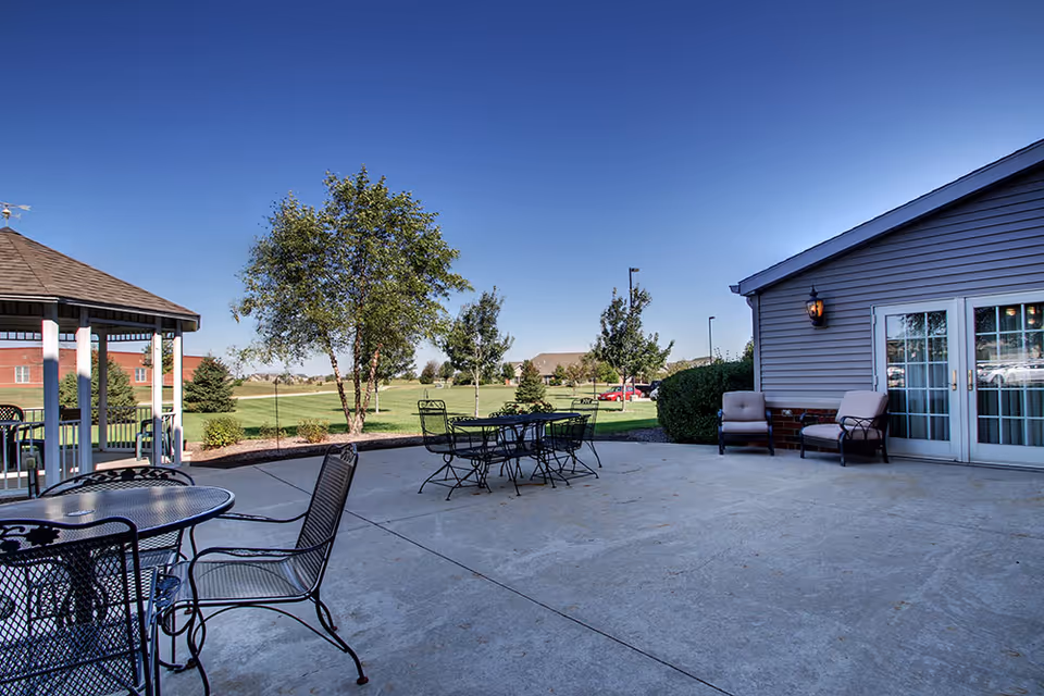Outdoor patio area at Evergreen Senior Living with metal tables and chairs, cushioned armchairs near a building with glass doors, a gazebo, and a grassy area with trees under a clear blue sky.