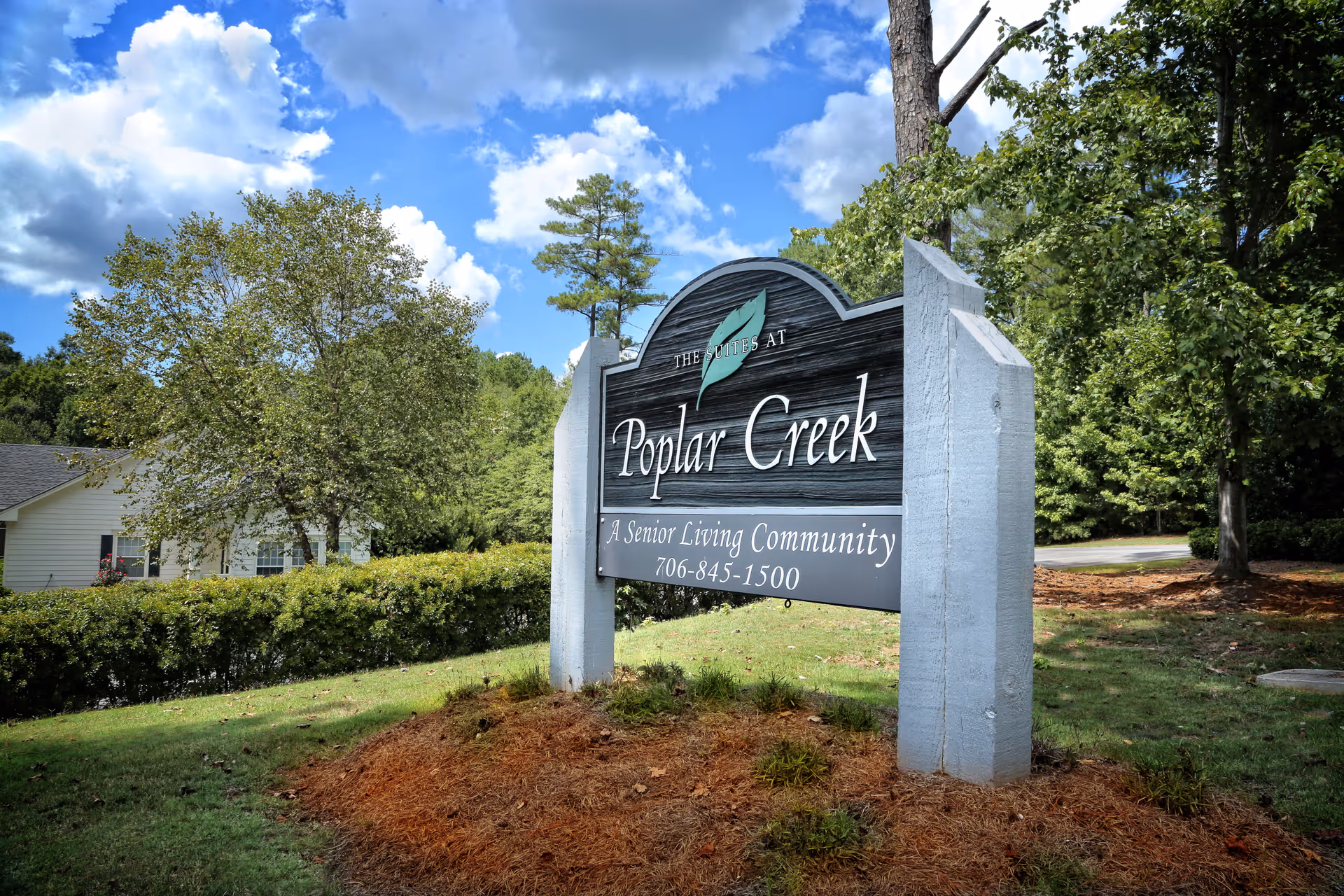 A landscaped entrance sign for Poplar Creek senior living community on a grassy lot with trees and a blue sky behind it.