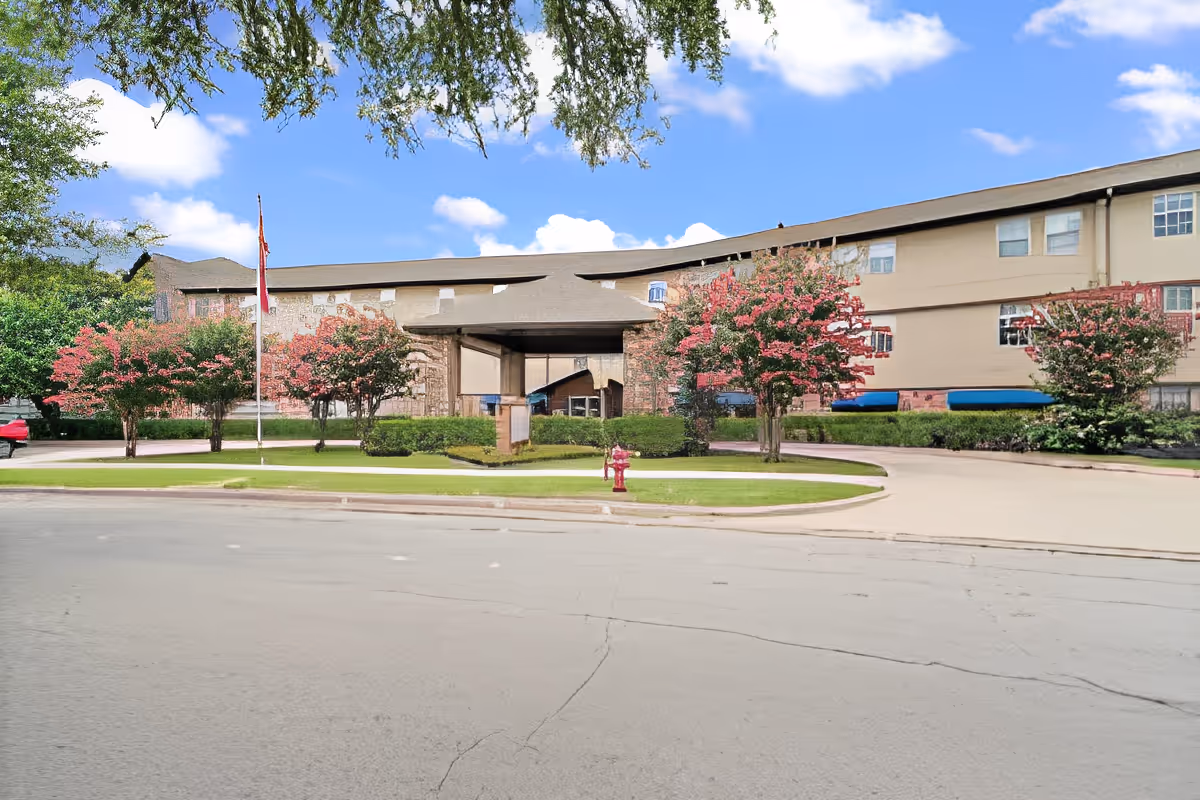 Front exterior of a senior living building with a covered entrance, circular driveway, landscaped trees, and a flagpole.