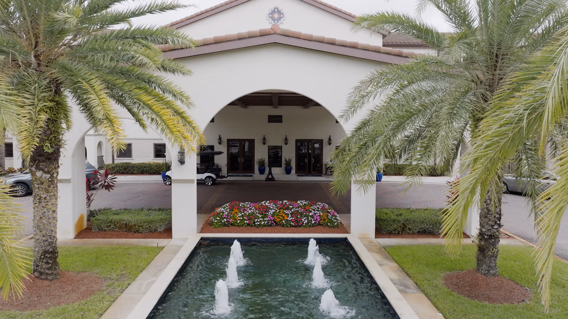 Entrance of The Arbor at Lake Worth facility featuring a covered driveway with an archway, a rectangular water fountain with four water jets in the foreground, palm trees on both sides, and a flower bed with colorful flowers near the building entrance.