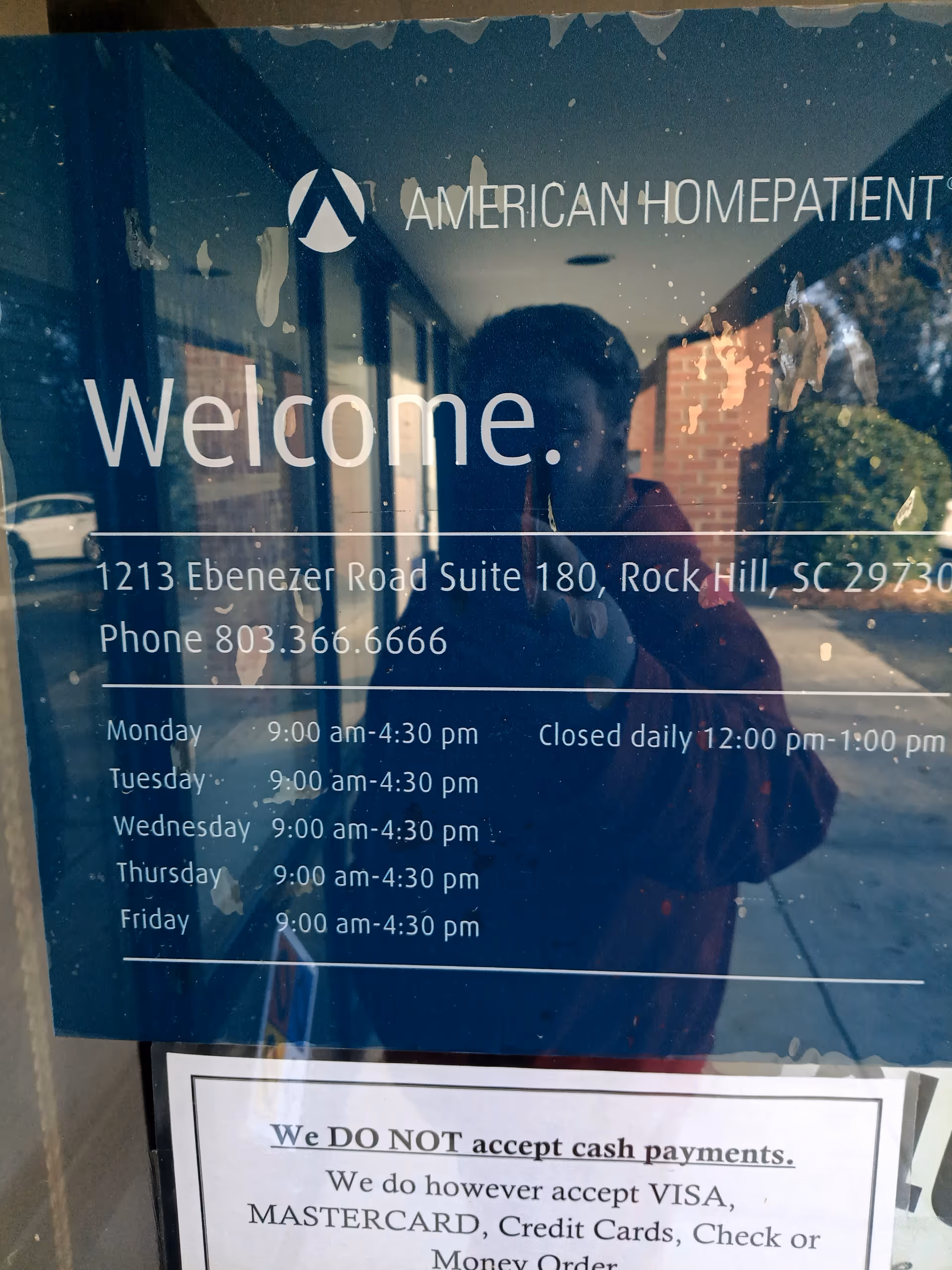 Reflective glass entrance door with Welcome sign, hours, and a person taking a photo.